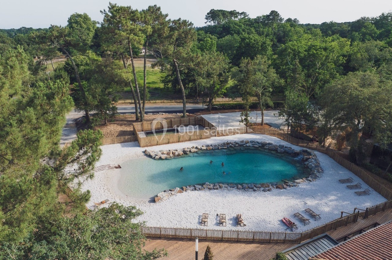 Vue aérienne d'une piscine naturelle entourée de sable blanc, de transats et de végétation arborée.
