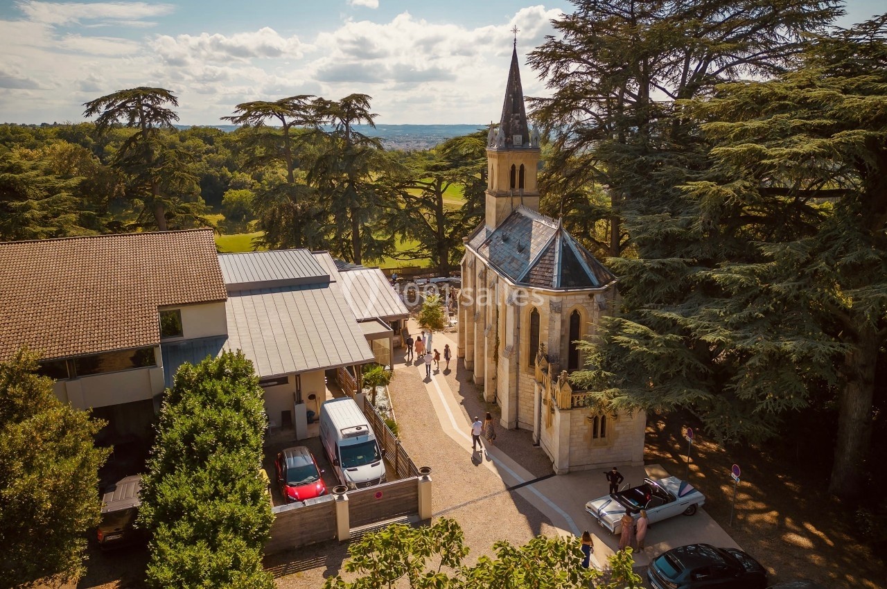 Vue aérienne d'une petite église entourée d'arbres, avec des bâtiments modernes et des voitures à proximité.