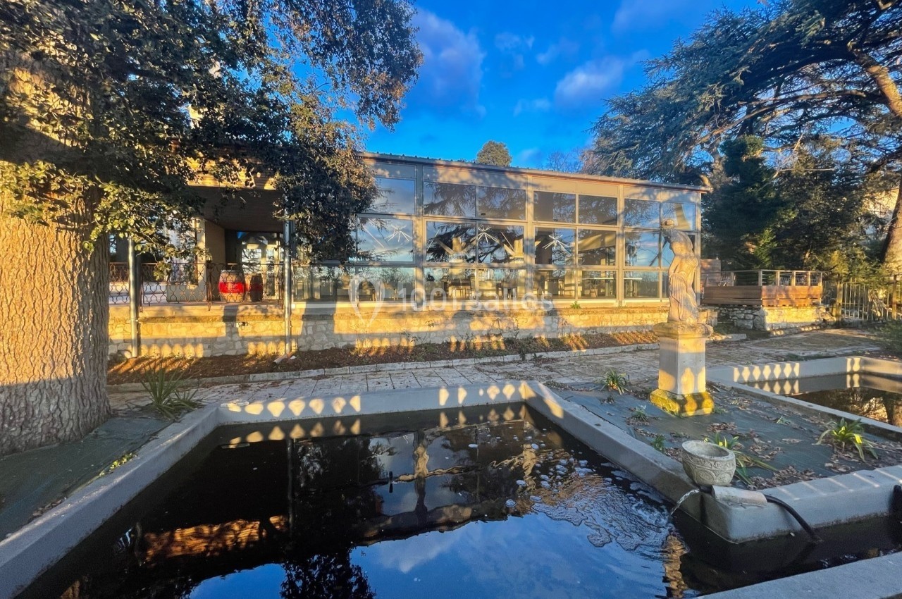 Vue d'un bâtiment moderne avec de grandes baies vitrées, entouré d'arbres et reflété dans un bassin au premier plan.