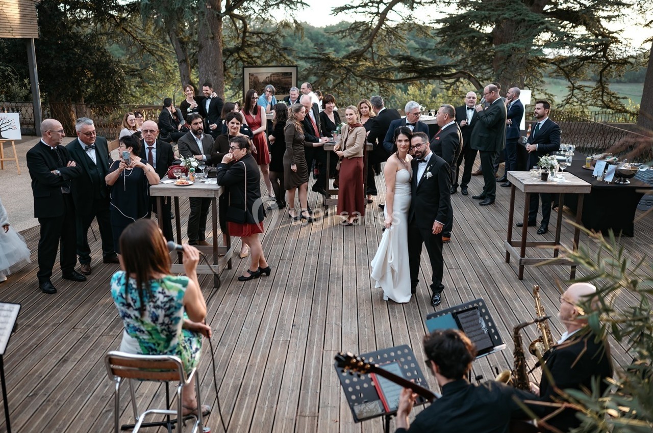 Un couple de mariés pose devant des invités rassemblés sur une terrasse en bois, avec des musiciens au premier plan.
