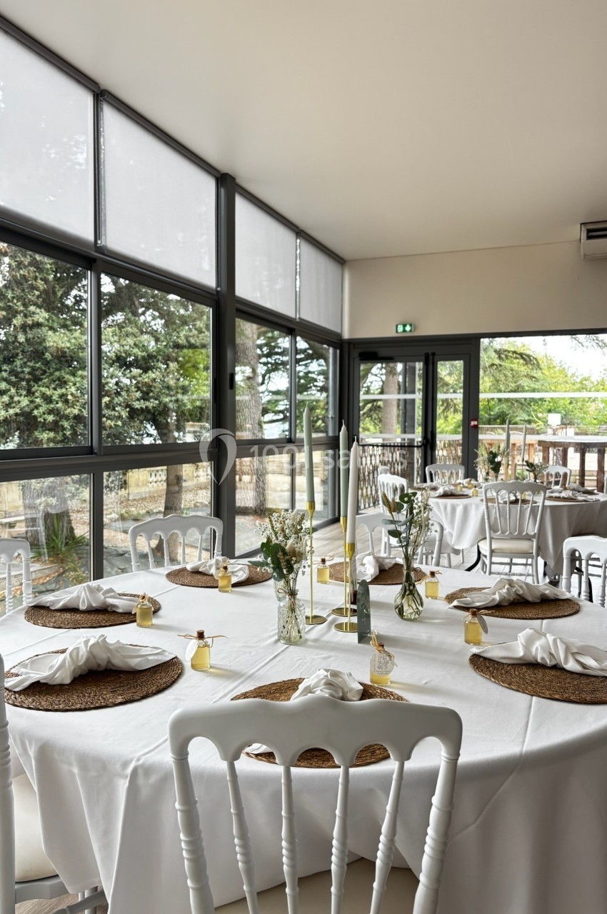 Salle lumineuse avec de grandes baies vitrées, tables rondes dressées avec nappes blanches, chaises blanches et décorations…