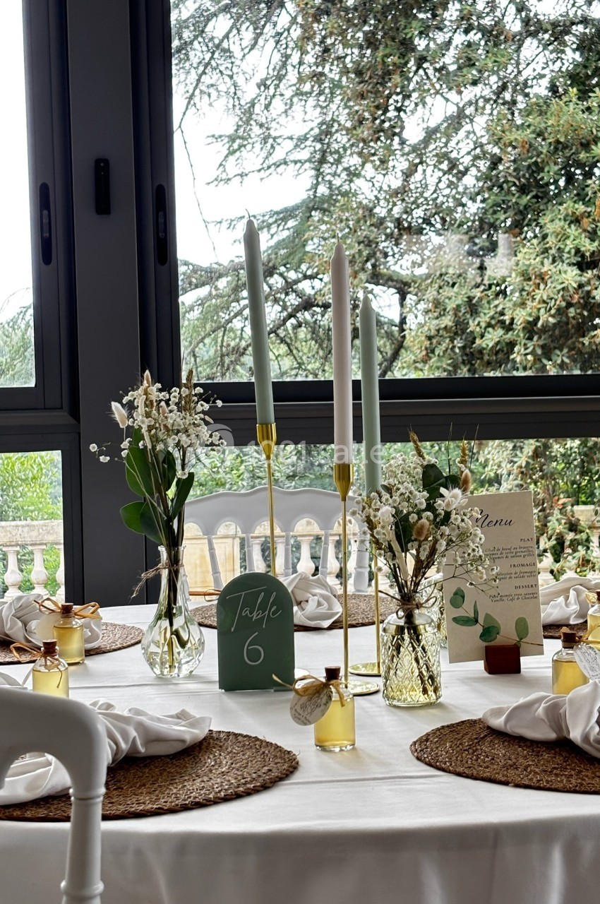 Table décorée pour un événement avec bougies, fleurs, menus et cadeaux, devant une baie vitrée donnant sur un jardin.