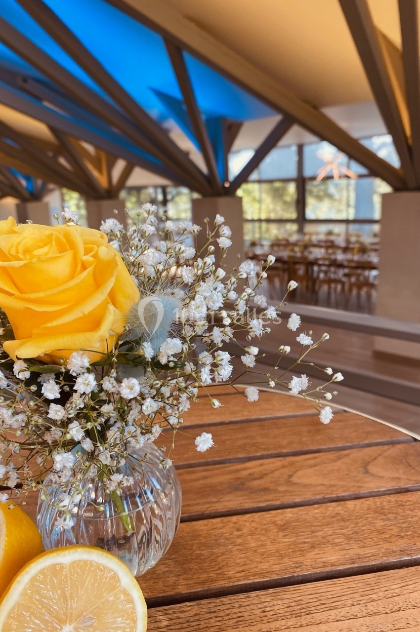 Vase en verre contenant une rose jaune et des gypsophiles, posé sur une table en bois dans un espace lumineux.