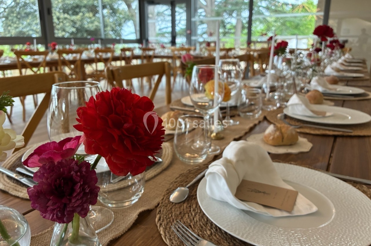 Table dressée avec élégance, ornée de fleurs rouges et violettes, avec vaisselle blanche et vue sur un extérieur verdoyant.