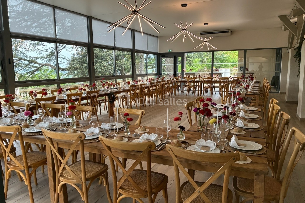 Salle lumineuse avec de grandes baies vitrées, tables en bois dressées avec vaisselle et décorations florales.
