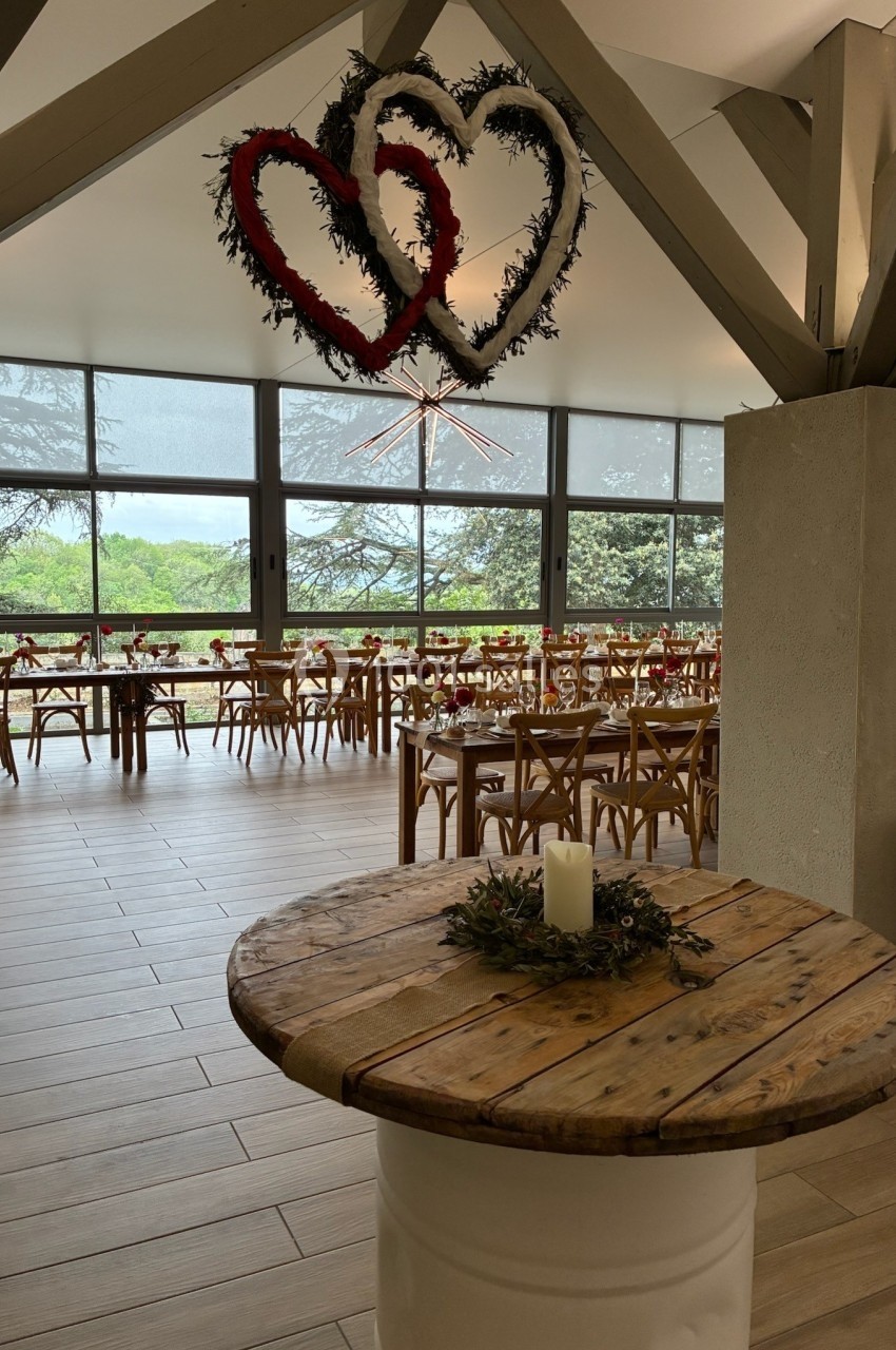 Salle de réception lumineuse avec tables décorées, deux cœurs suspendus et une bougie sur une table en bois.
