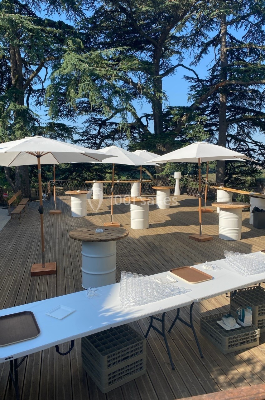 Terrasse en bois aménagée pour un événement avec tables hautes, parasols blancs et vaisselle disposée.