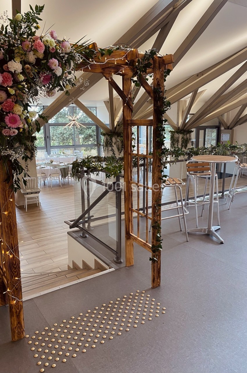 Arche en bois décorée de fleurs et de guirlandes lumineuses dans une salle lumineuse avec des tables hautes et des chaises.