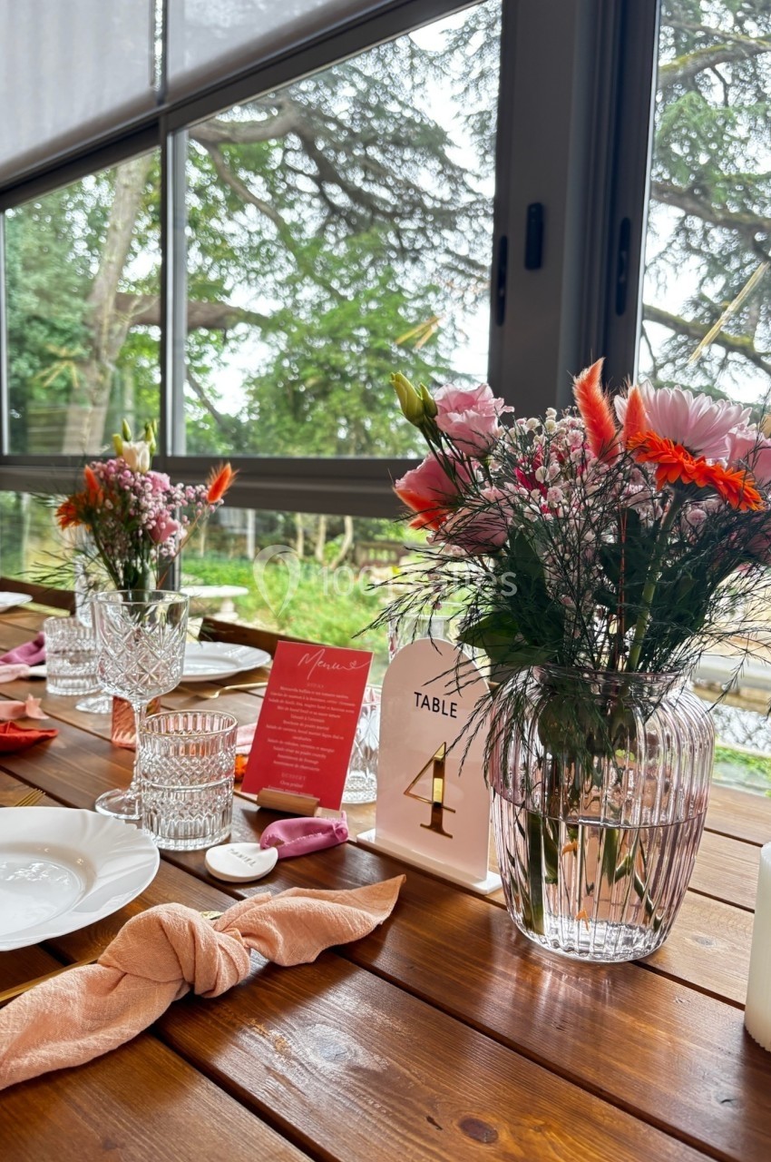 Table en bois décorée avec des fleurs colorées, des serviettes roses et des cartes, près d'une grande baie vitrée donnant…