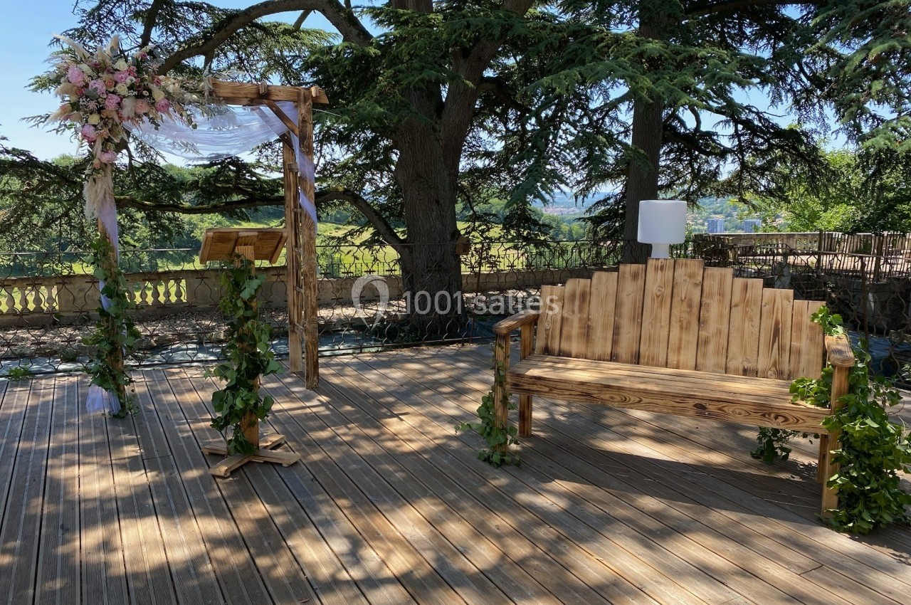 Arche en bois décorée de fleurs et banc assorti sur une terrasse en bois, entourée d'arbres et de verdure.