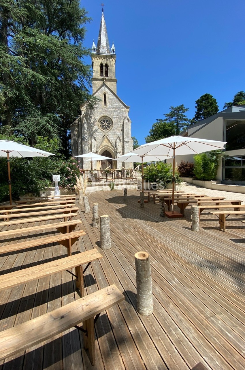 Terrasse en bois avec bancs et parasols blancs, face à une église en pierre entourée de verdure par temps ensoleillé.