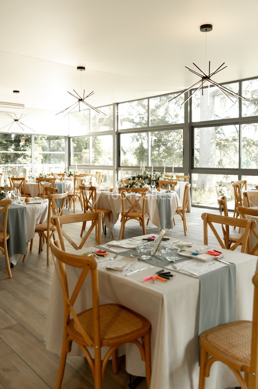 Salle lumineuse avec de grandes baies vitrées, tables dressées avec nappes blanches et chaises en bois clair.