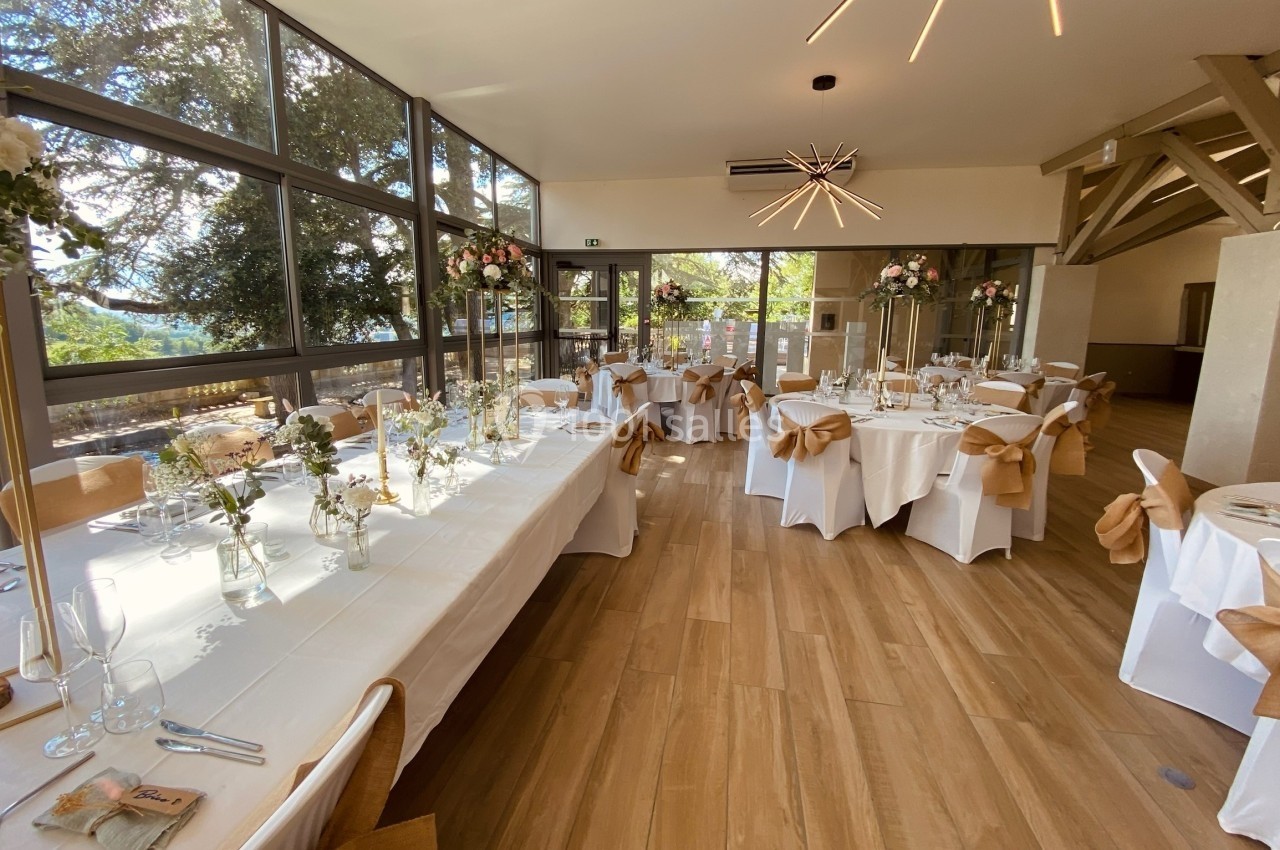 Salle de réception lumineuse avec tables décorées de nappes blanches, fleurs et nœuds en tissu beige.