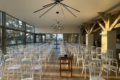 Salle en pierre avec parquet, tables dressées pour un repas, grandes fenêtres et éclairage suspendu.