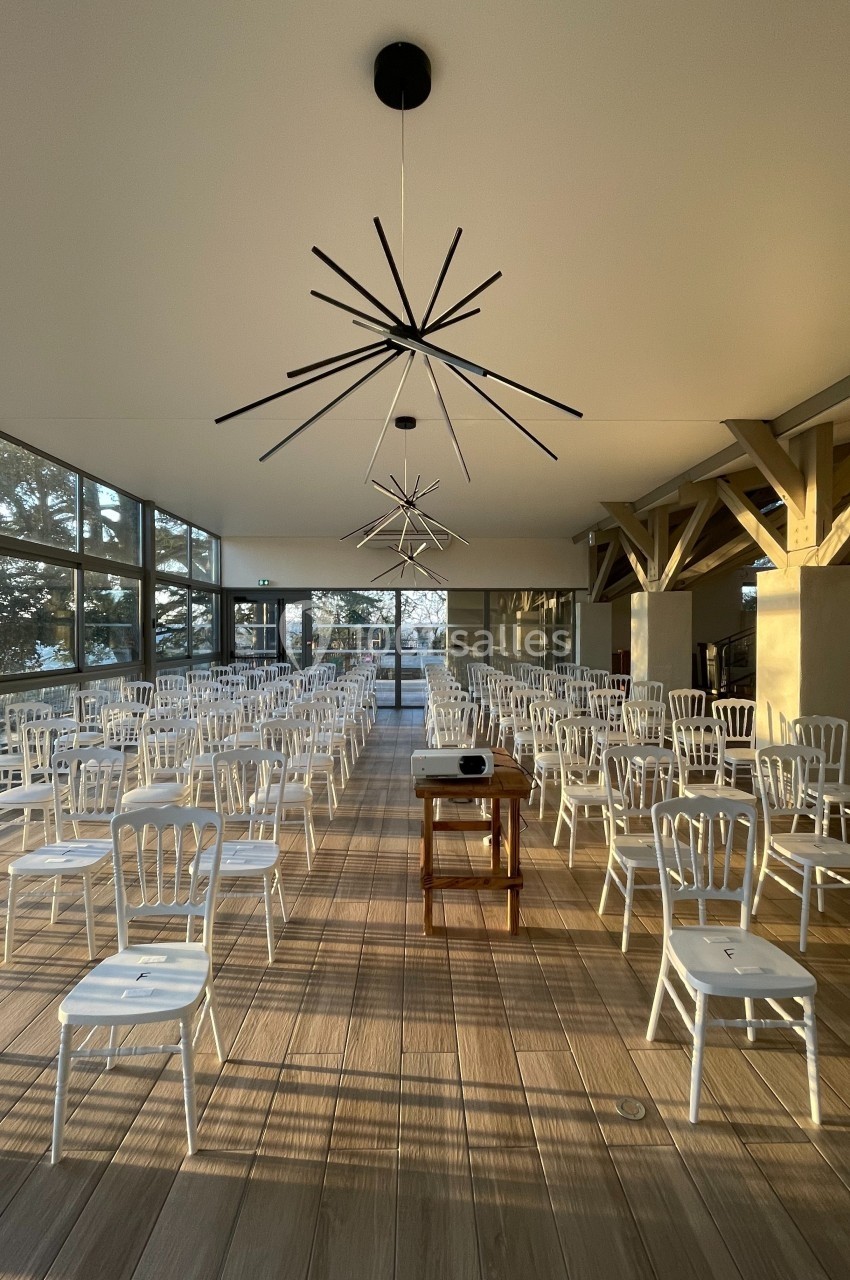 Salle lumineuse avec des chaises blanches alignées, un projecteur sur une table et de grandes baies vitrées.
