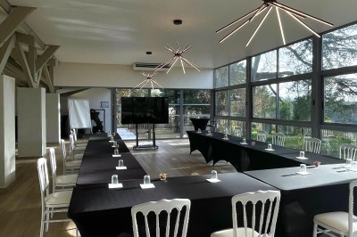 Salle en pierre avec parquet, tables dressées pour un repas, grandes fenêtres et éclairage suspendu.