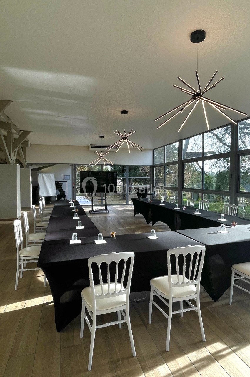 Salle de réunion lumineuse avec grandes baies vitrées, tables en U couvertes de nappes noires et chaises blanches.