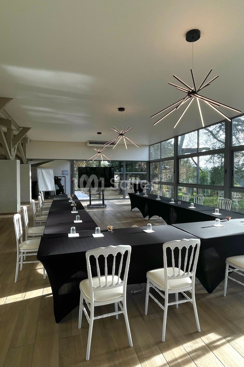 Salle de réunion lumineuse avec grandes baies vitrées, tables en U couvertes de nappes noires et chaises blanches.