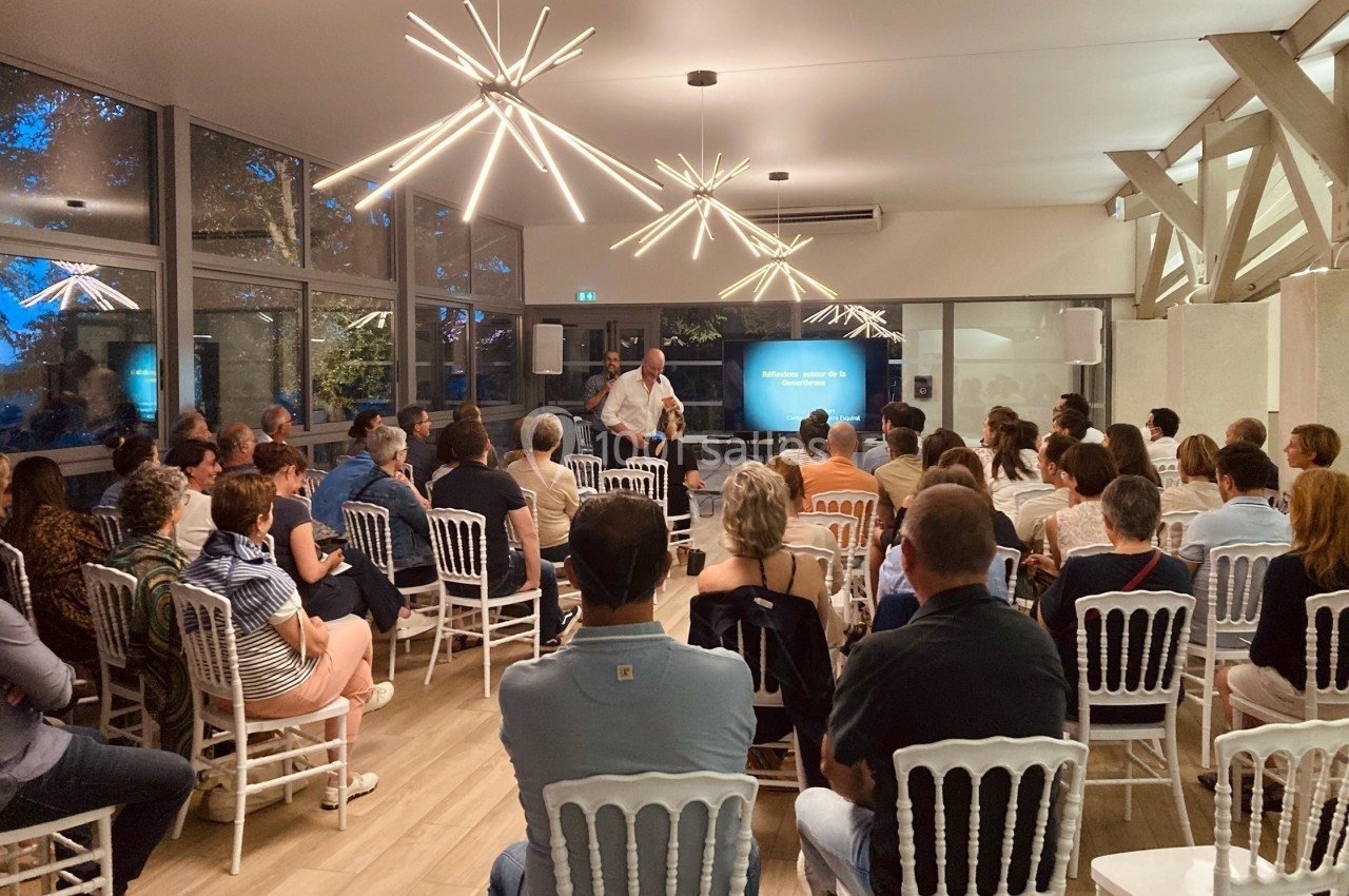 Un conférencier s'adresse à un public assis dans une salle lumineuse avec des chaises blanches et des éclairages modernes.