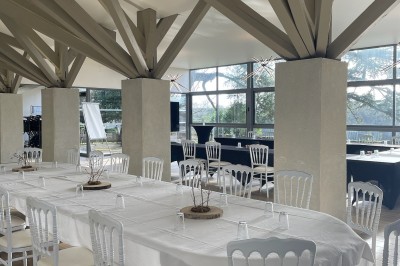 Salle en pierre avec parquet, tables dressées pour un repas, grandes fenêtres et éclairage suspendu.