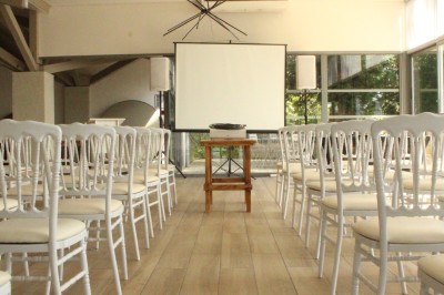 Salle en pierre avec parquet, tables dressées pour un repas, grandes fenêtres et éclairage suspendu.