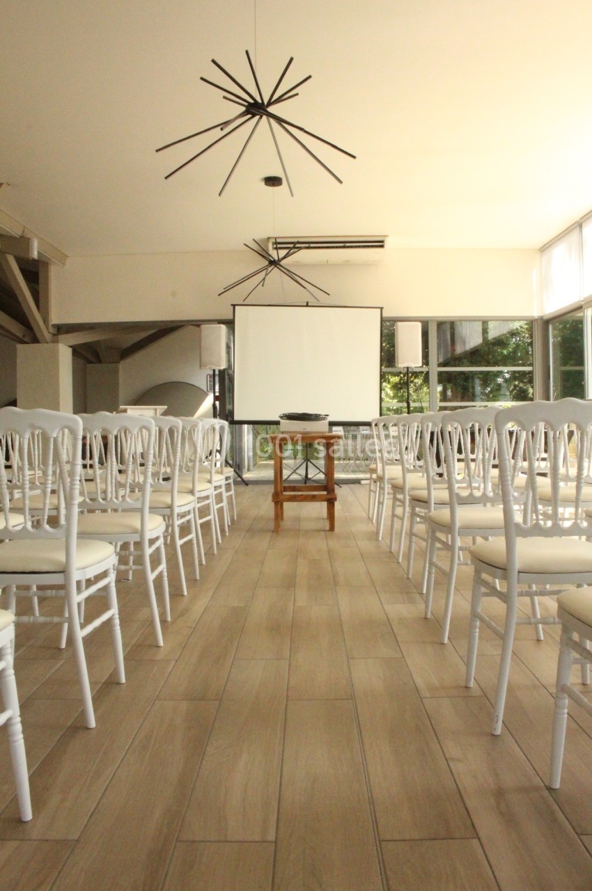 Salle lumineuse aménagée pour une présentation, avec des rangées de chaises blanches et un écran de projection.