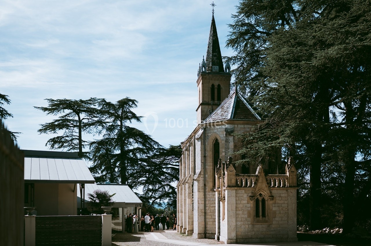 Petite église en pierre avec un clocher, entourée d'arbres et de bâtiments modernes sous un ciel dégagé.