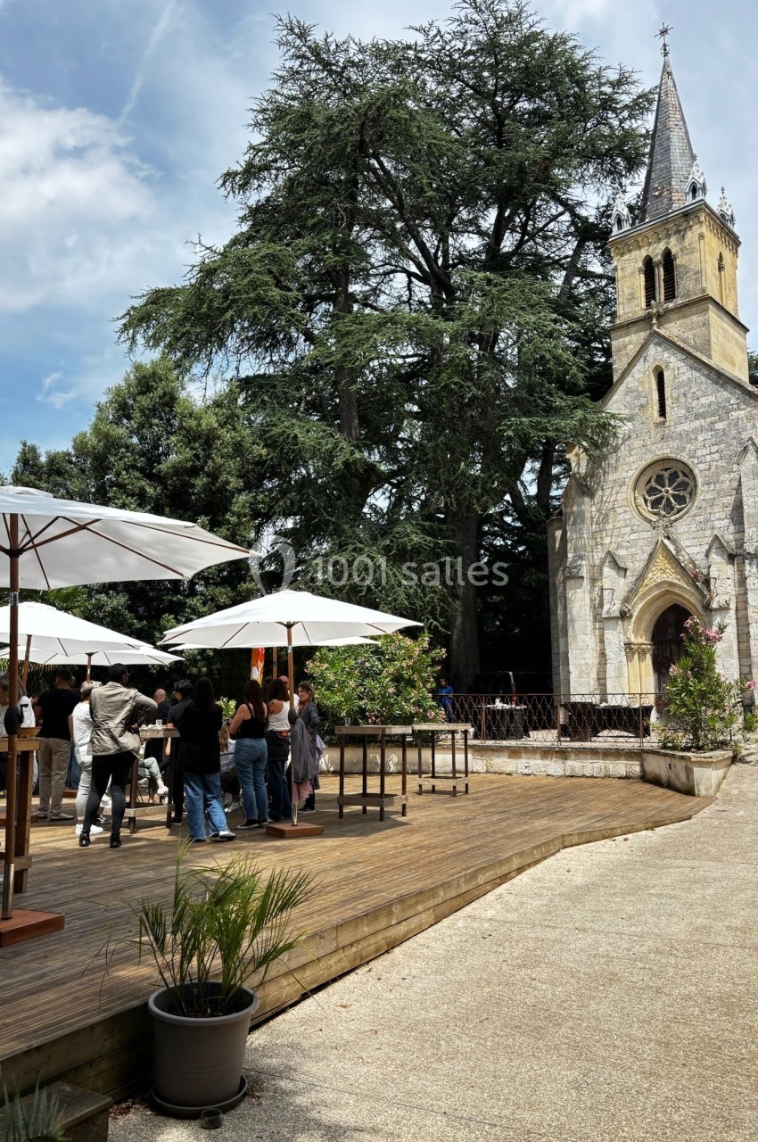 Terrasse en bois avec parasols et visiteurs près d'une petite église en pierre entourée d'arbres.