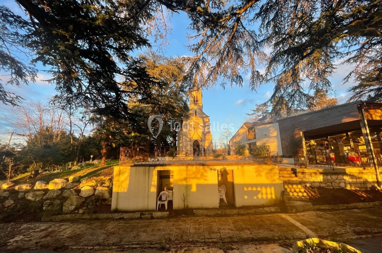 Vue d'une petite église en pierre éclairée par le soleil couchant, entourée d'arbres et d'un bâtiment moderne.