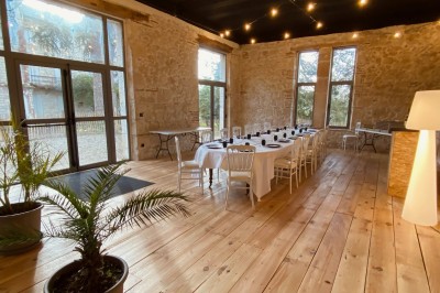 Salle en pierre avec parquet, tables dressées pour un repas, grandes fenêtres et éclairage suspendu.