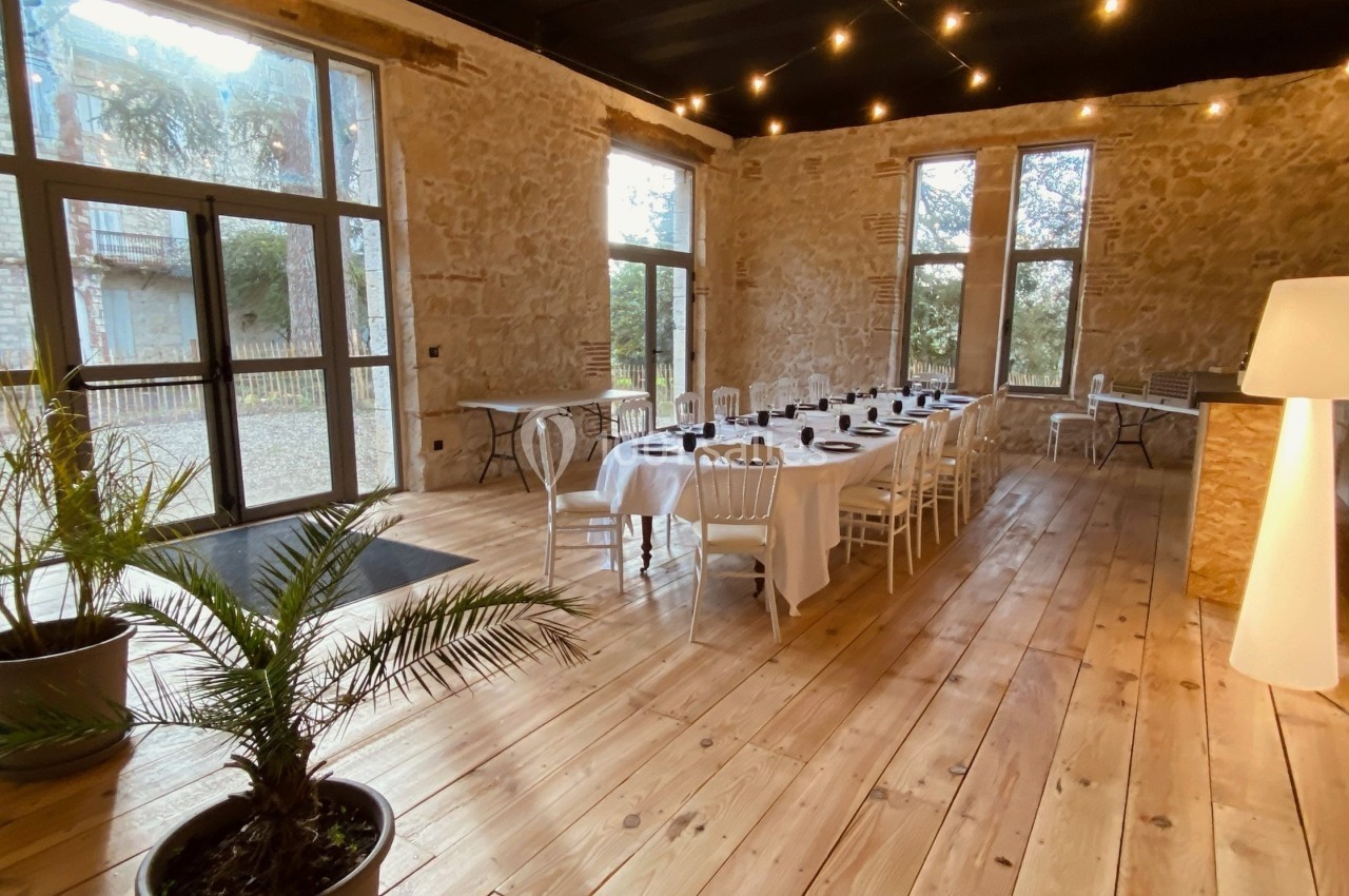 Salle en pierre avec parquet, tables dressées pour un repas, grandes fenêtres et éclairage suspendu.