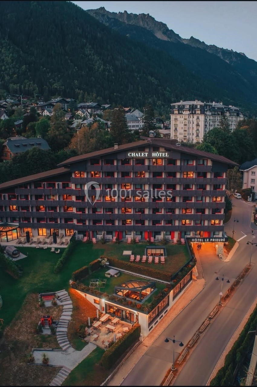 Vue aérienne d'un chalet-hôtel éclairé au crépuscule, entouré de montagnes et de bâtiments dans un cadre alpin.