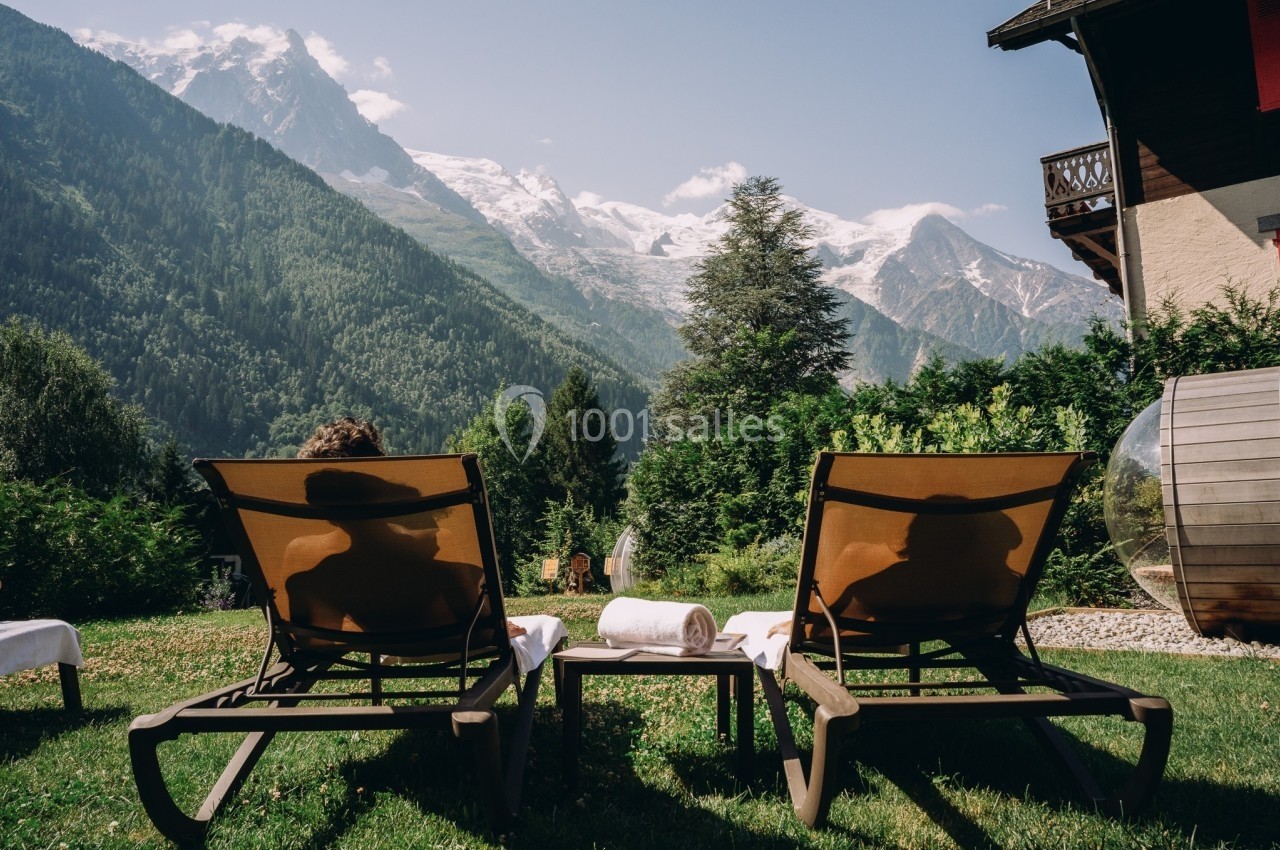 Deux personnes allongées sur des transats dans un jardin, face à des montagnes enneigées et entourées de verdure.