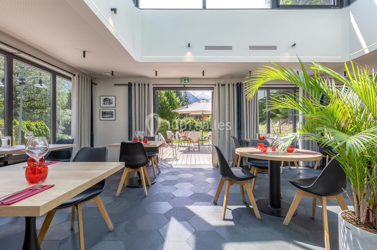 Salle de restaurant lumineuse avec tables en bois, chaises noires, grandes baies vitrées et vue sur une terrasse extérieure.