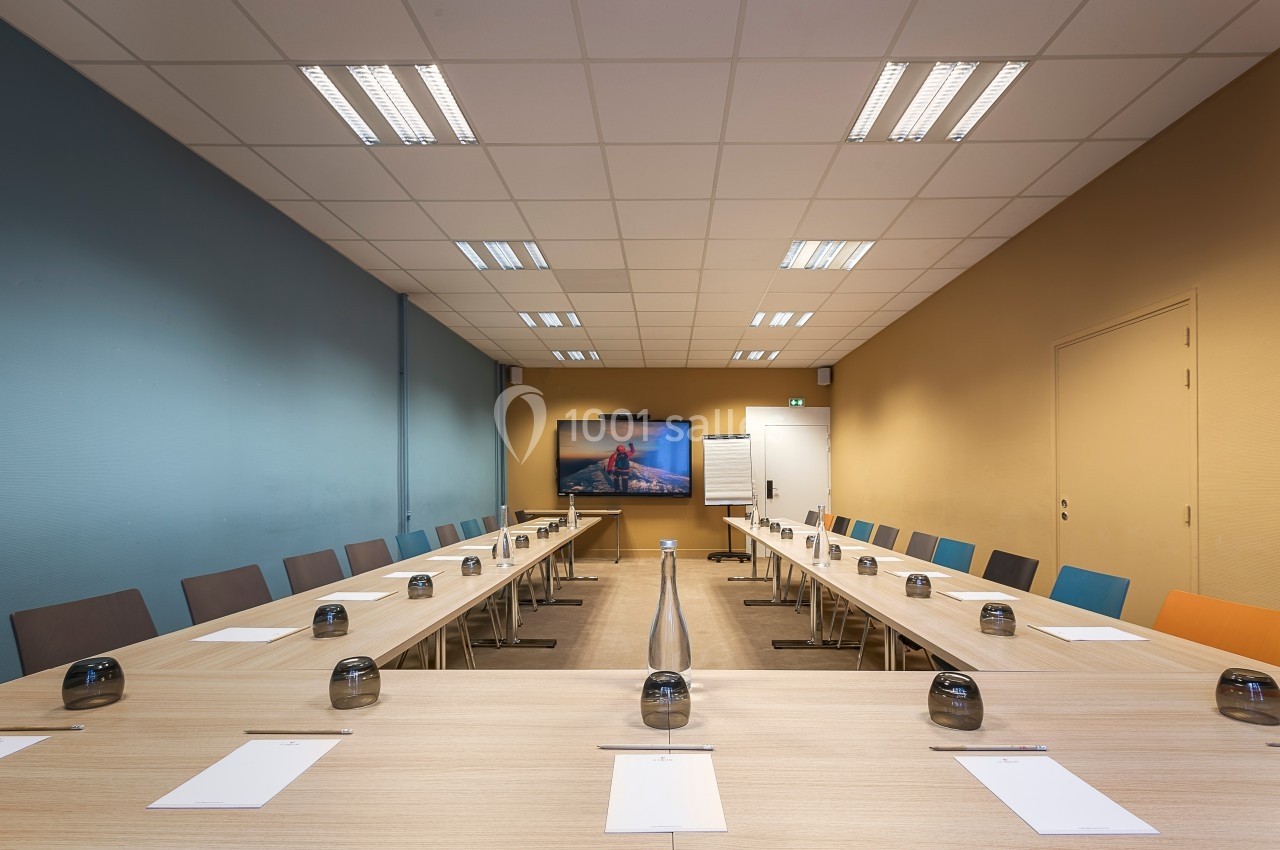 Salle de réunion lumineuse avec une grande table en U, des chaises colorées et un écran mural allumé.