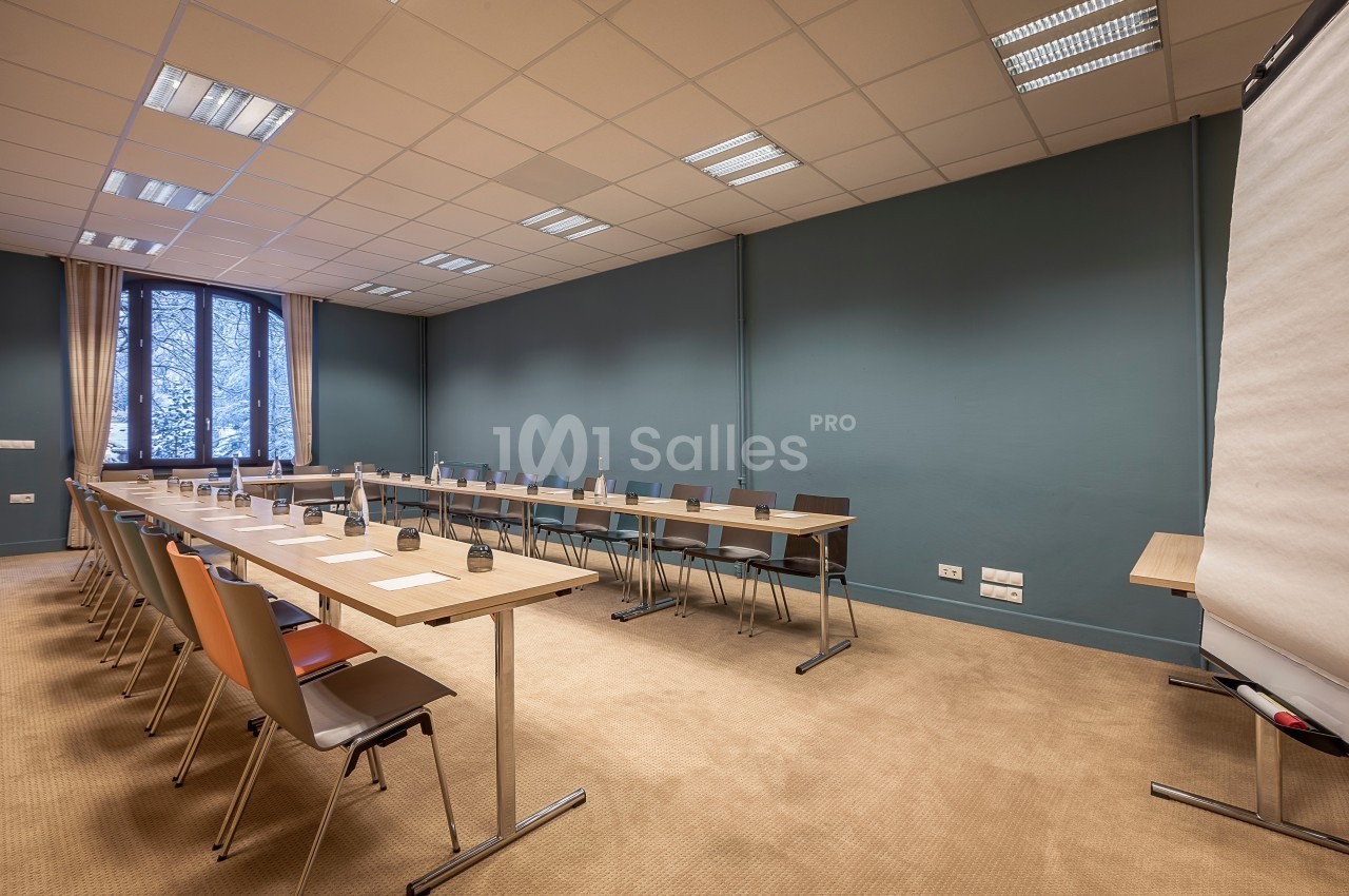 Salle de réunion avec tables disposées en U, chaises alignées, tableau de conférence et grande fenêtre.