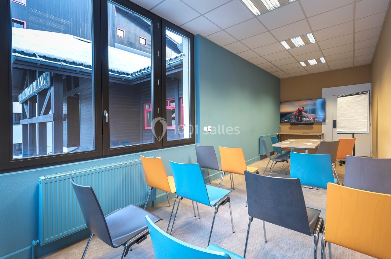 Salle de réunion lumineuse avec chaises colorées, grande fenêtre donnant sur l'extérieur et écran au fond.