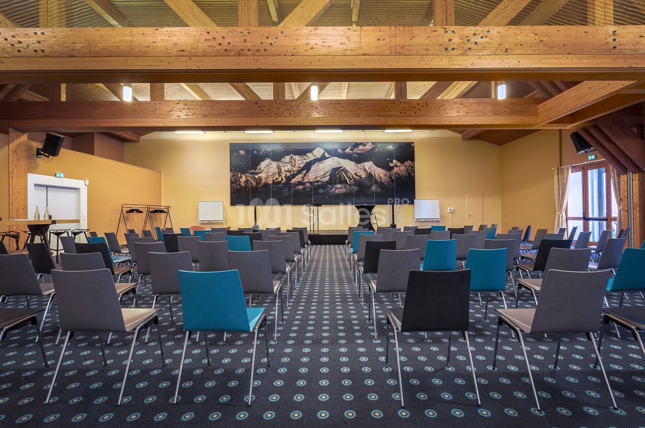 Salle de conférence vide avec des rangées de chaises, un podium et un décor mural représentant des montagnes.
