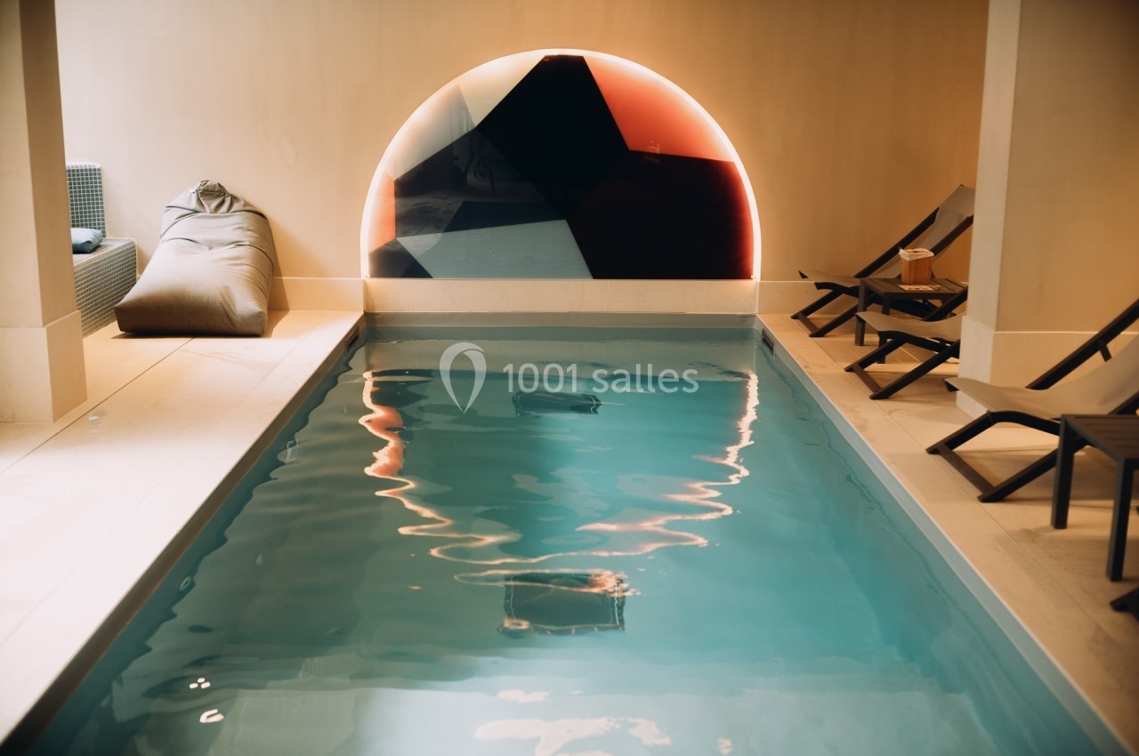 Piscine intérieure avec eau claire, entourée de chaises longues et éclairée par une lumière murale colorée.