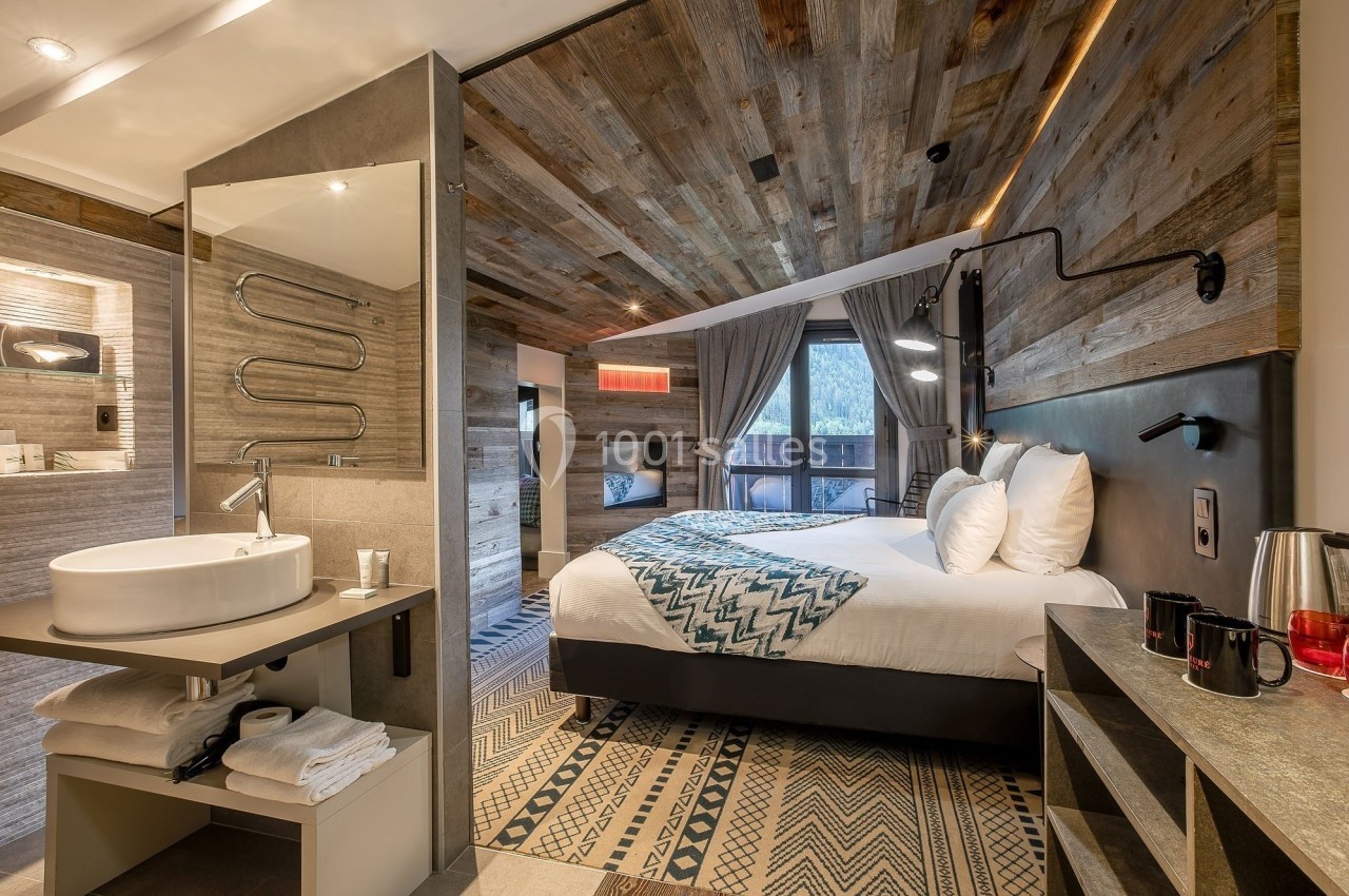 Chambre d'hôtel moderne avec lit double, décor en bois, salle de bain ouverte et vue sur l'extérieur par une baie vitrée.
