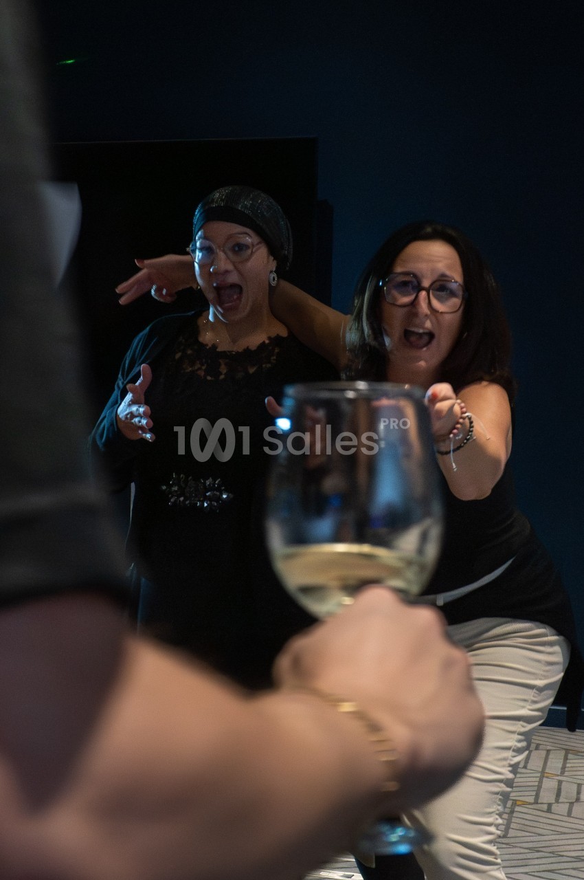 Deux femmes réagissent avec surprise et amusement en direction d'un verre de vin blanc tendu vers elles.