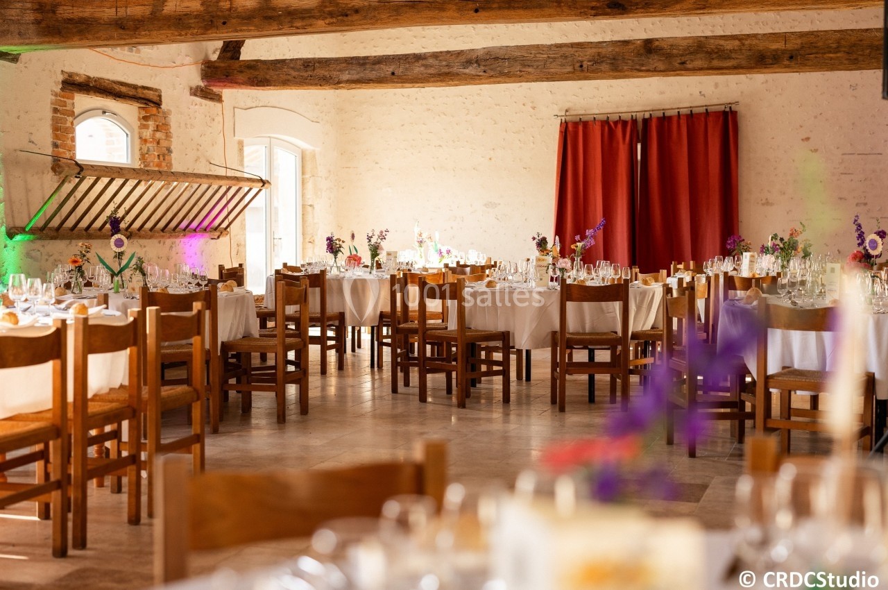Salle de réception lumineuse avec tables décorées de fleurs et chaises en bois, poutres apparentes au plafond.