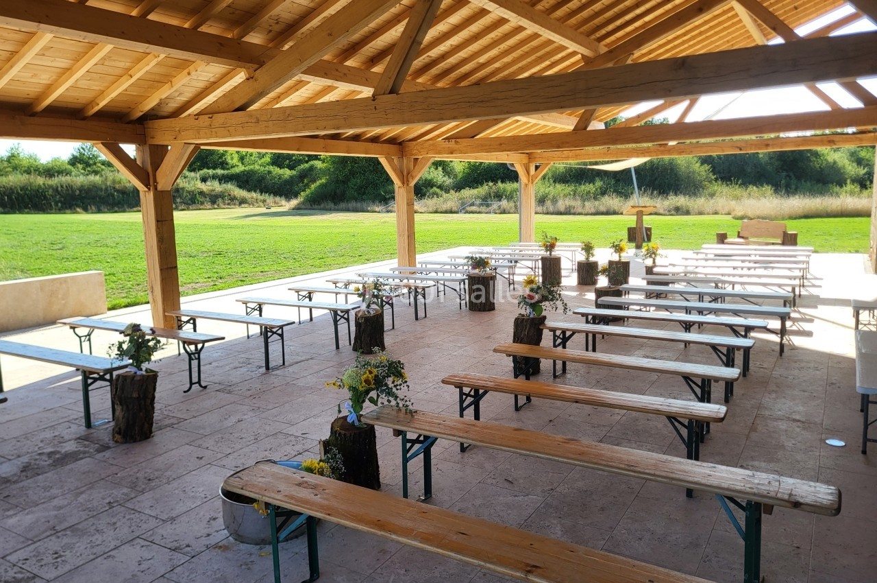 Tables et bancs en bois disposés sous une pergola, décorés de fleurs, avec un espace vert en arrière-plan.