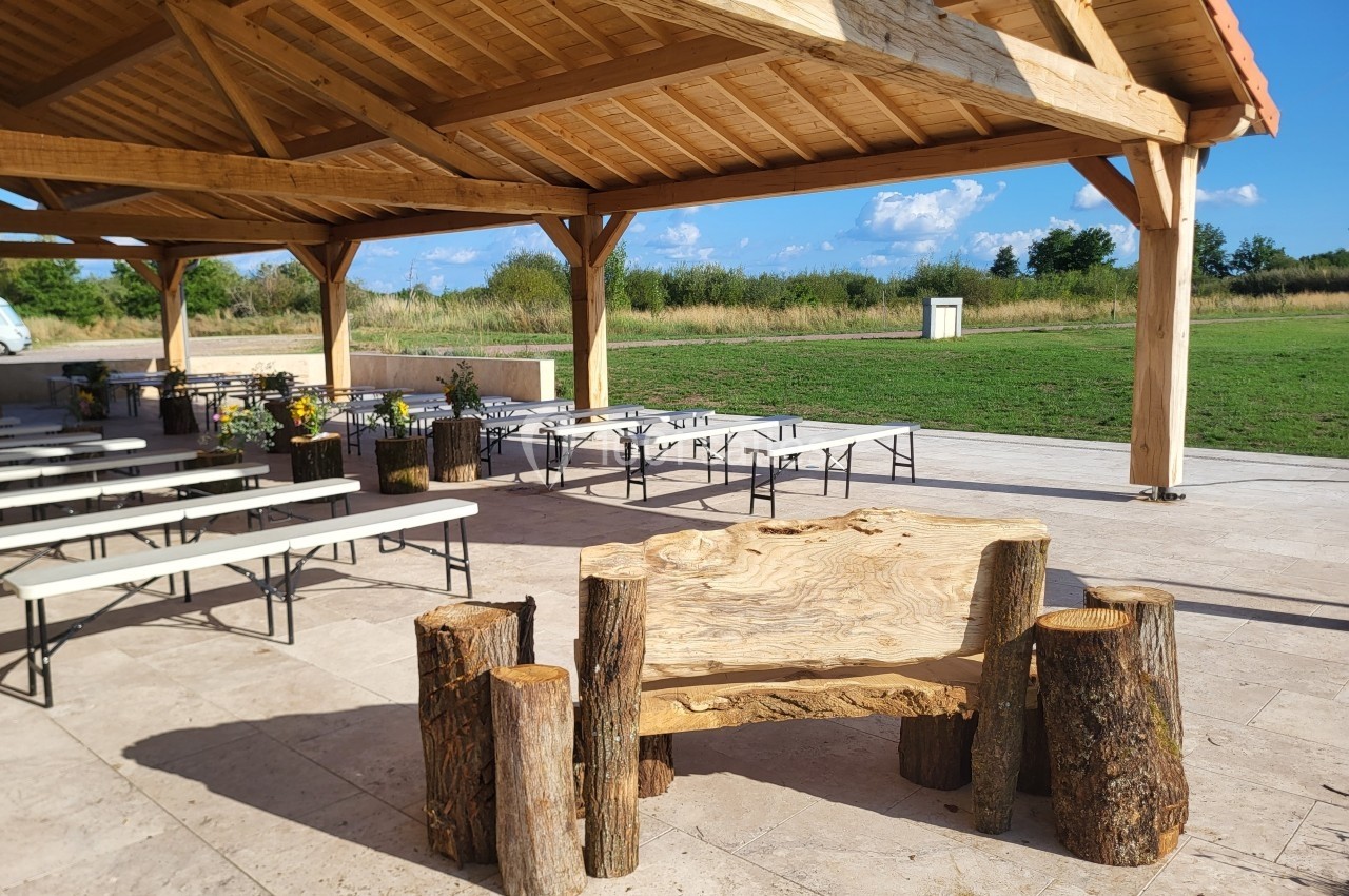 Abri en bois avec tables et bancs sur une terrasse en pierre, entouré d'un espace vert en arrière-plan.