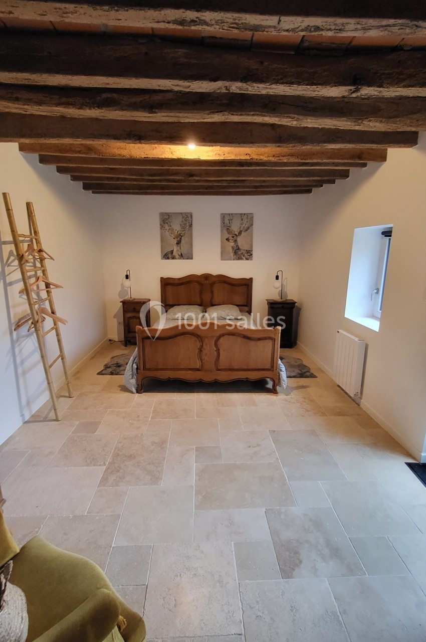 Chambre avec lit en bois sculpté, poutres apparentes, murs blancs, sol en pierre et décoration minimaliste.