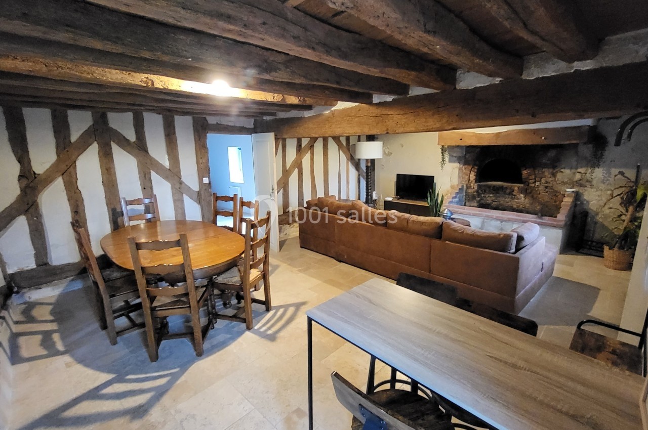Salle de séjour rustique avec poutres apparentes, table en bois, canapé, cheminée en pierre et lumière naturelle.