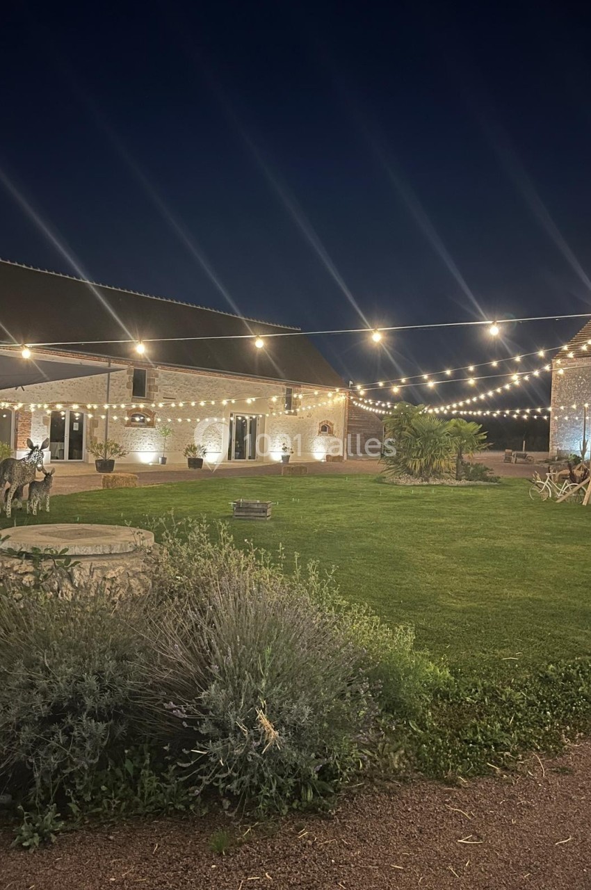 Cour extérieure illuminée par des guirlandes lumineuses, entourée de bâtiments en pierre et d'un jardin verdoyant.
