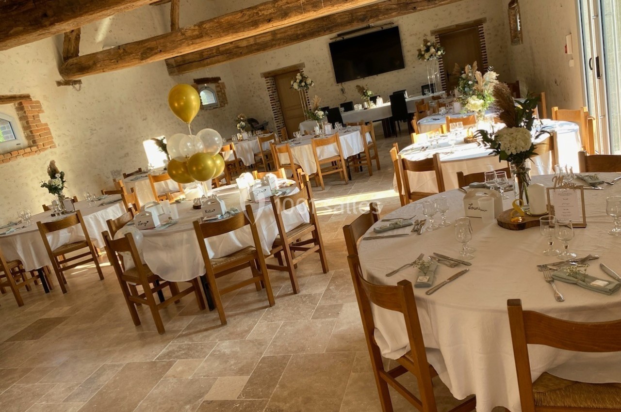 Salle de réception lumineuse avec tables rondes dressées, décorations florales et ballons dorés.