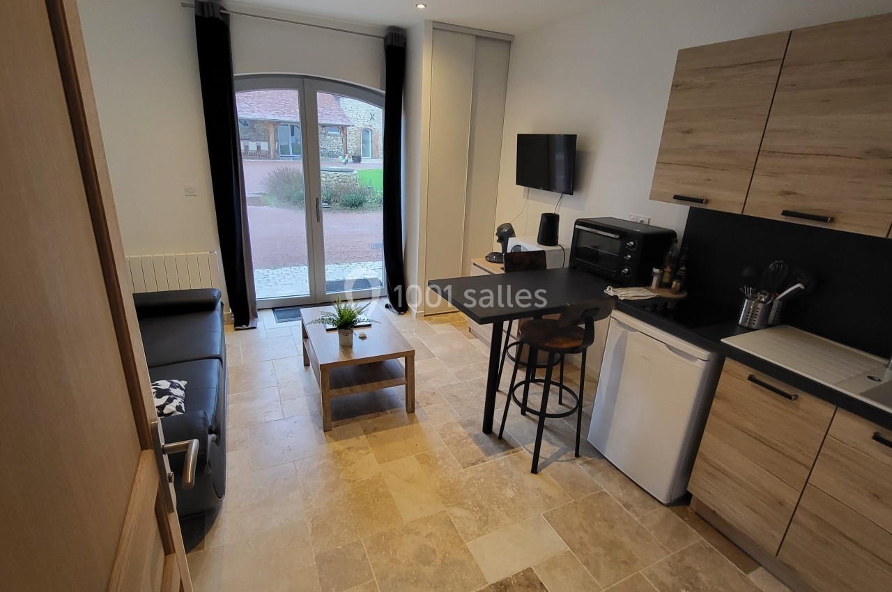 Studio moderne avec coin cuisine équipé, table haute, canapé, et grande porte vitrée donnant sur une cour extérieure.