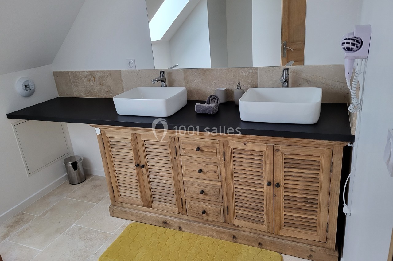 Meuble de salle de bain en bois avec deux vasques blanches, miroir, sèche-cheveux mural et tapis jaune au sol.